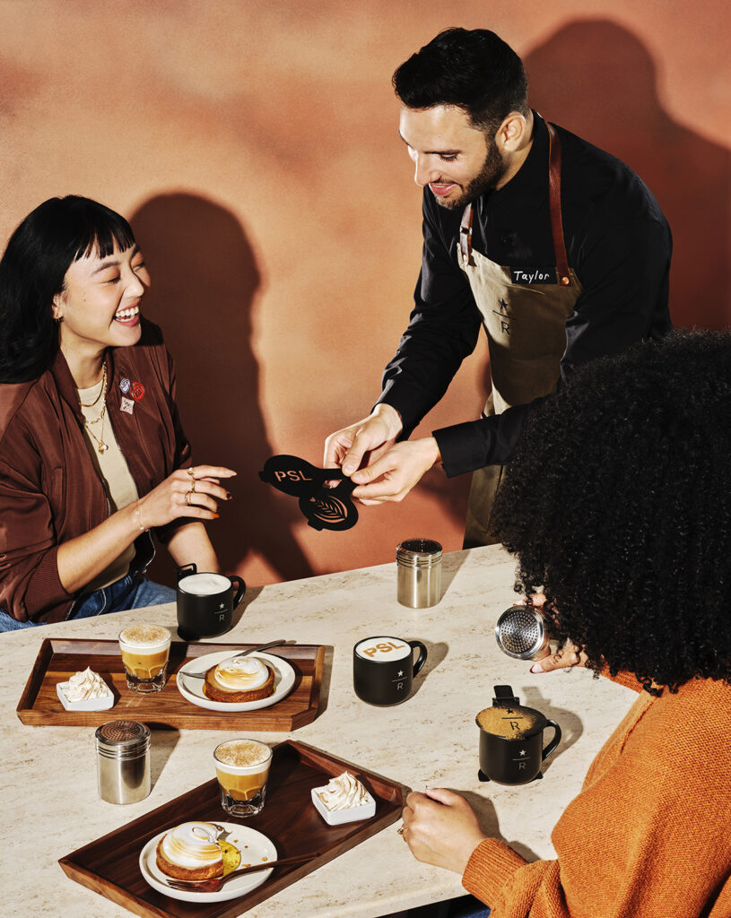 A barista serves a woman at a table with coffee drinks and desserts. Two mugs are present, one with "PSL" and another with "R." The setting has warm tones and a cozy atmosphere.