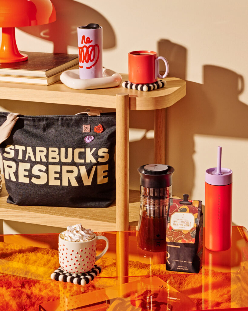 A vibrant display featuring a red and white polka dot mug with whipped cream, a purple travel mug, and a black coffee maker. A Starbucks Reserve bag and a bright orange table complete the scene.