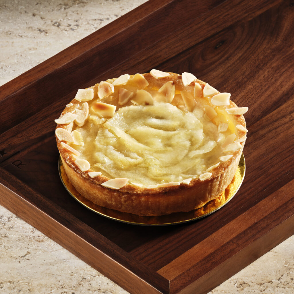A round cake with a golden crust, topped with a smooth, creamy layer and garnished with almond slivers, sits on a wooden tray.