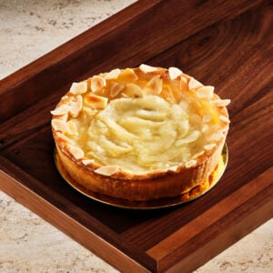A round cake with a golden crust, topped with a smooth, creamy layer and garnished with almond slivers, sits on a wooden tray.