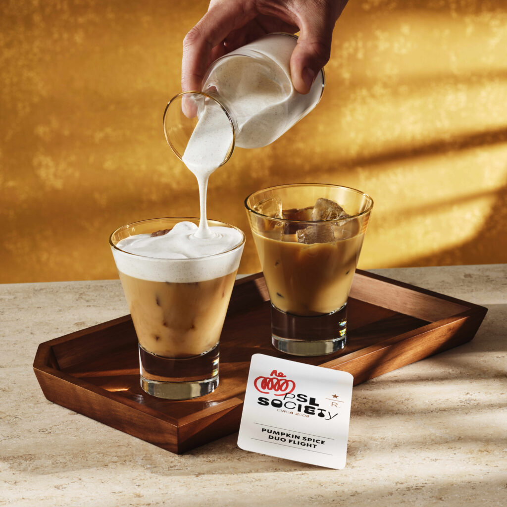 A hand pours frothy cream into a glass of iced coffee, while another glass with ice sits nearby. A card labeled "Pumpkin Spice Duo Flight" rests on a wooden tray. The background features a warm, golden hue.