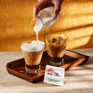 A hand pours frothy cream into a glass of iced coffee, while another glass with ice sits nearby. A card labeled "Pumpkin Spice Duo Flight" rests on a wooden tray. The background features a warm, golden hue.