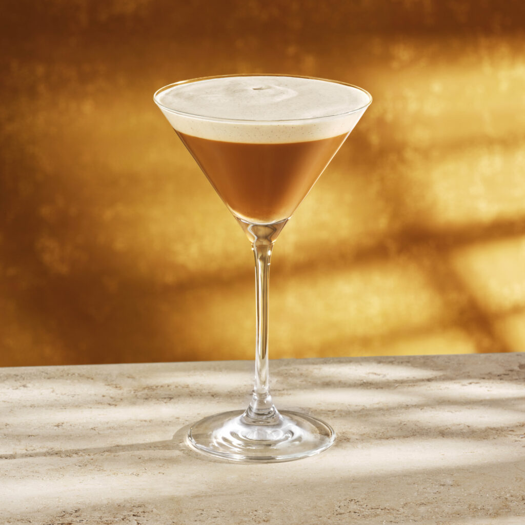 A sleek martini glass holds a layered drink with a rich brown base and a frothy cream top, set against a warm golden background.