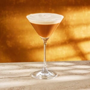 A sleek martini glass holds a layered drink with a rich brown base and a frothy cream top, set against a warm golden background.