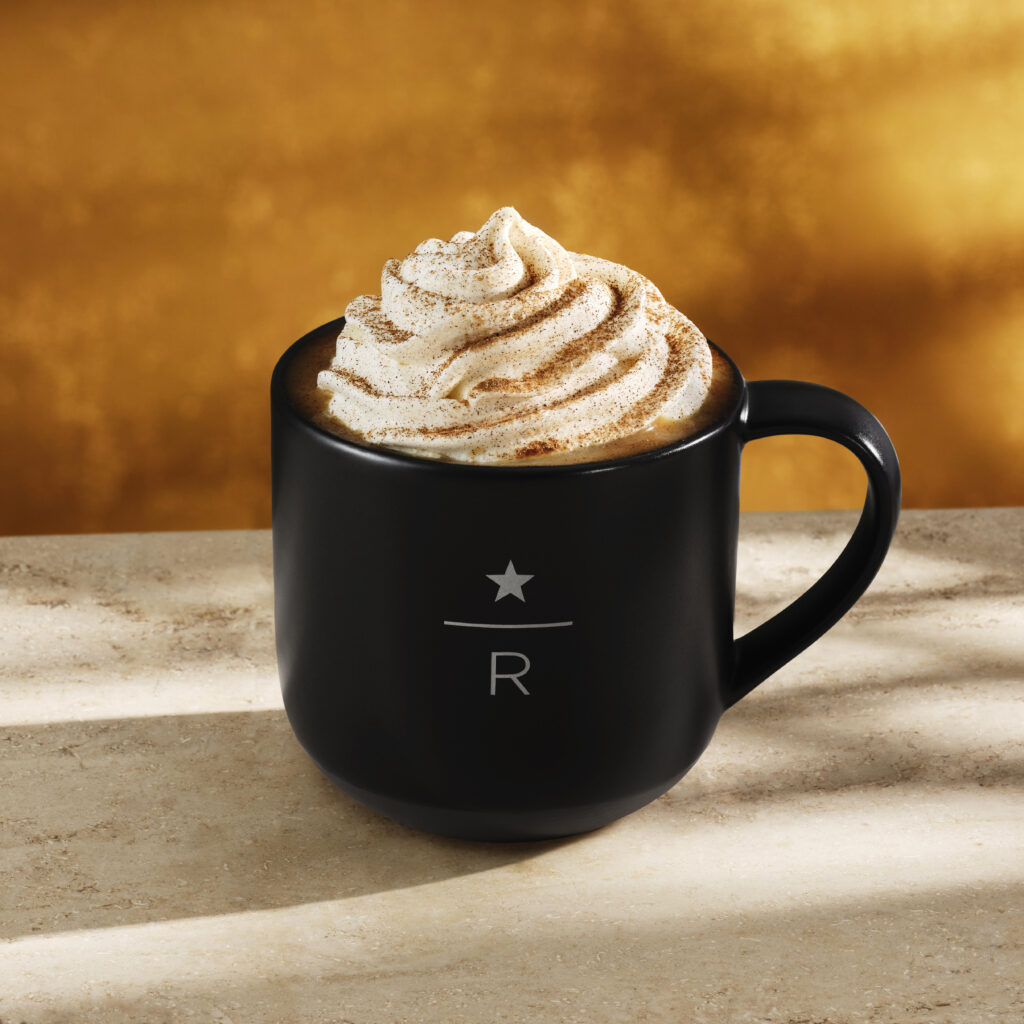 A black mug filled with coffee topped with whipped cream and a sprinkle of cinnamon, set on a light stone surface with a warm, golden background.