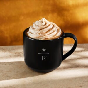 A black mug filled with coffee topped with whipped cream and a sprinkle of cinnamon, set on a light stone surface with a warm, golden background.