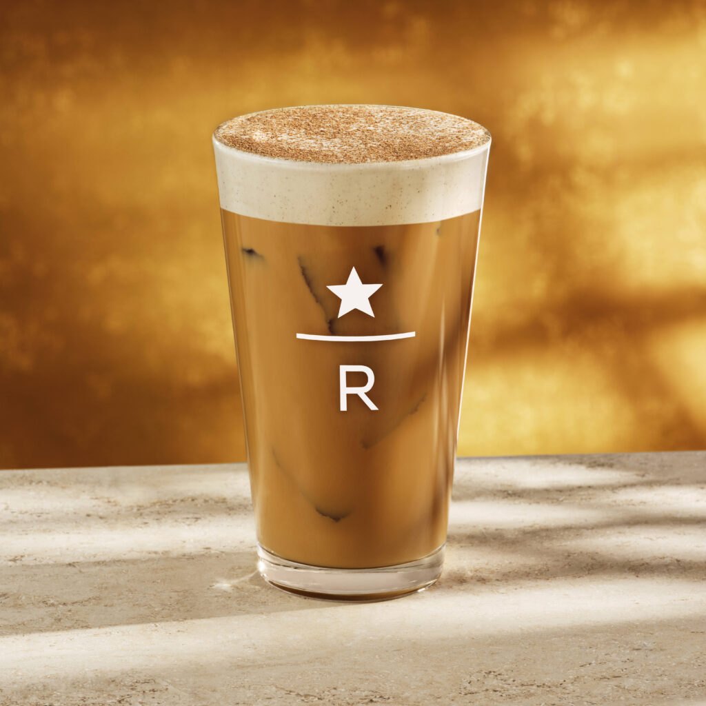 A tall glass filled with iced coffee topped with frothy cream and a sprinkle of cinnamon, set against a warm golden background.