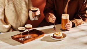 Two hands hold cocktails, one with a frothy top, while a dessert with a toasted meringue sits on a plate. A wooden tray holds another cocktail. A tall drink with foam is also present on the table.