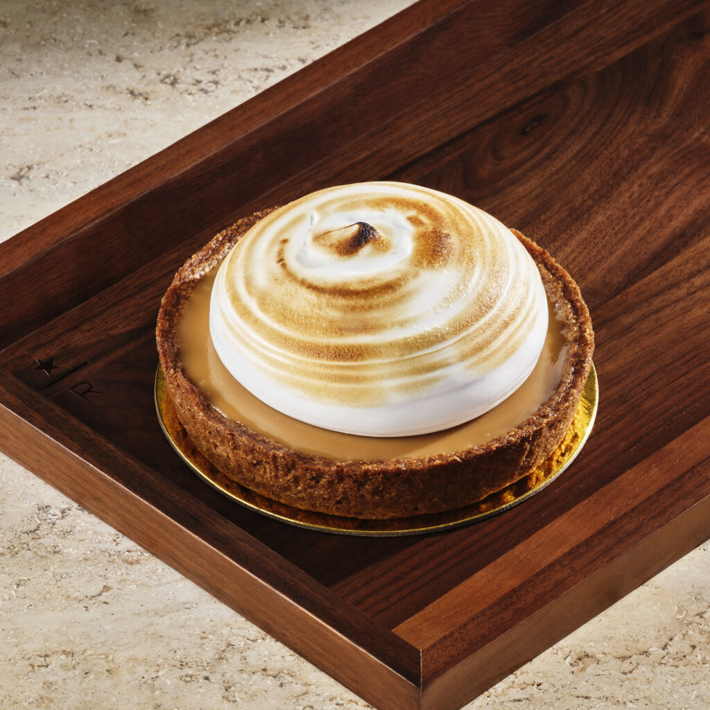 A round dessert with a golden-brown crust, topped with a fluffy, toasted meringue swirl. It sits on a wooden tray.