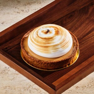 A round dessert with a golden-brown crust, topped with a fluffy, toasted meringue swirl. It sits on a wooden tray.