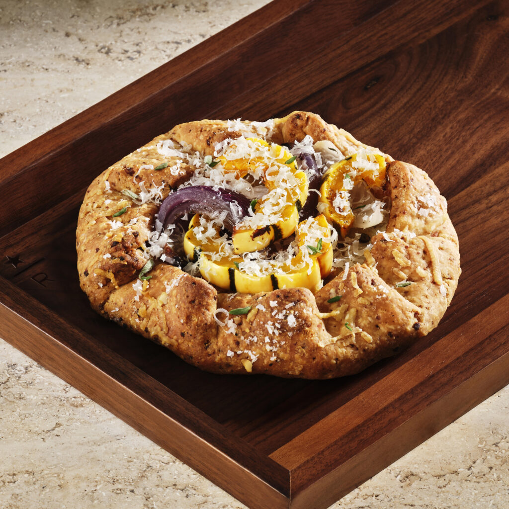 A rustic galette features a golden-brown crust filled with colorful roasted vegetables, including squash and onions, topped with grated cheese. It's presented on a wooden tray.