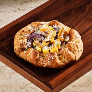 A rustic galette features a golden-brown crust filled with colorful roasted vegetables, including squash and onions, topped with grated cheese. It's presented on a wooden tray.