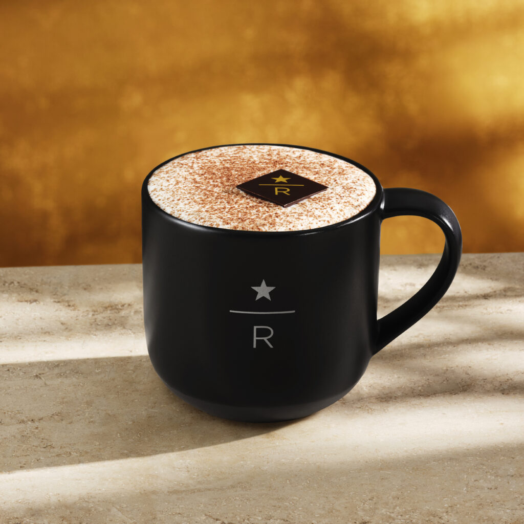 A black coffee mug sits on a light stone surface, filled with a frothy beverage topped with cocoa powder and a chocolate square. The background features a warm golden hue.