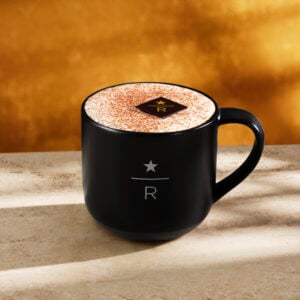A black coffee mug sits on a light stone surface, filled with a frothy beverage topped with cocoa powder and a chocolate square. The background features a warm golden hue.