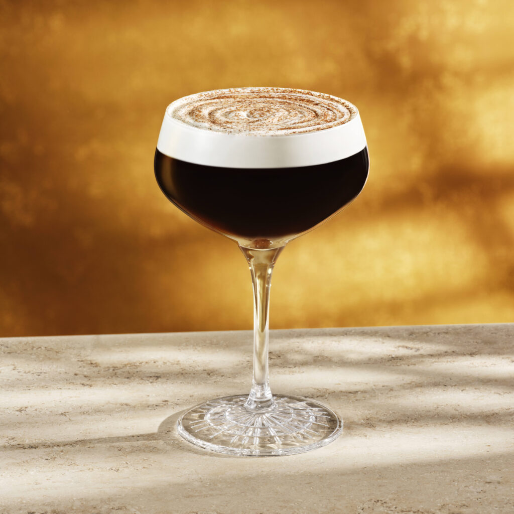 A stylish cocktail glass filled with dark liquid, topped with a frothy layer and a dusting of cocoa powder, sits on a light-colored surface. The background features a warm, golden hue.