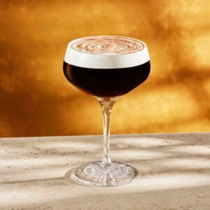 A stylish cocktail glass filled with dark liquid, topped with a frothy layer and a dusting of cocoa powder, sits on a light-colored surface. The background features a warm, golden hue.