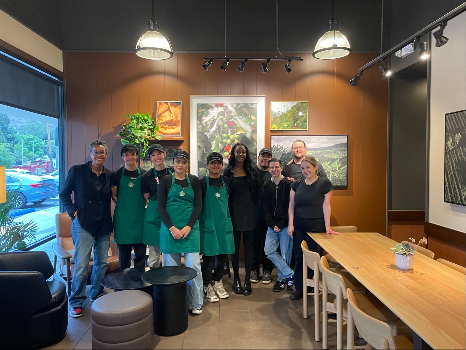 A group of eight people stands together in a coffee shop, wearing green aprons. The interior features wooden accents, plants, and artwork.