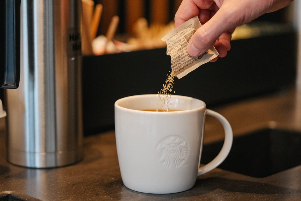 A hand is pouring sugar from a packet into a white Starbucks mug filled with coffee. The background features a dark surface and blurred items.