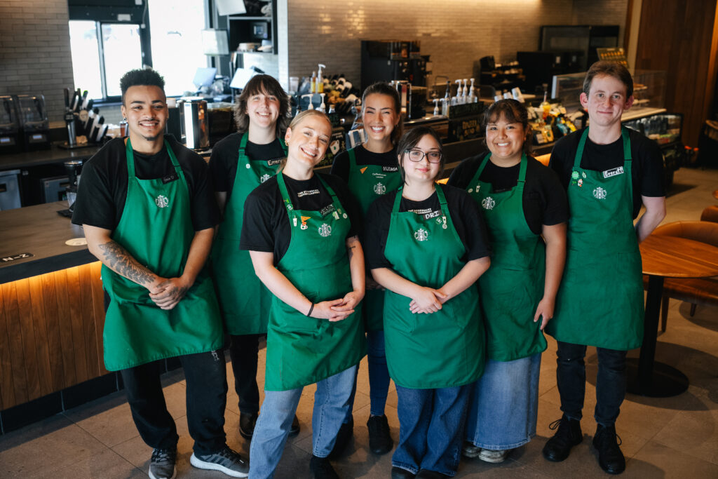 A group of seven baristas stands together, all wearing green aprons over black shirts. They are smiling and posing in a coffee shop setting, with equipment and a counter visible in the background.