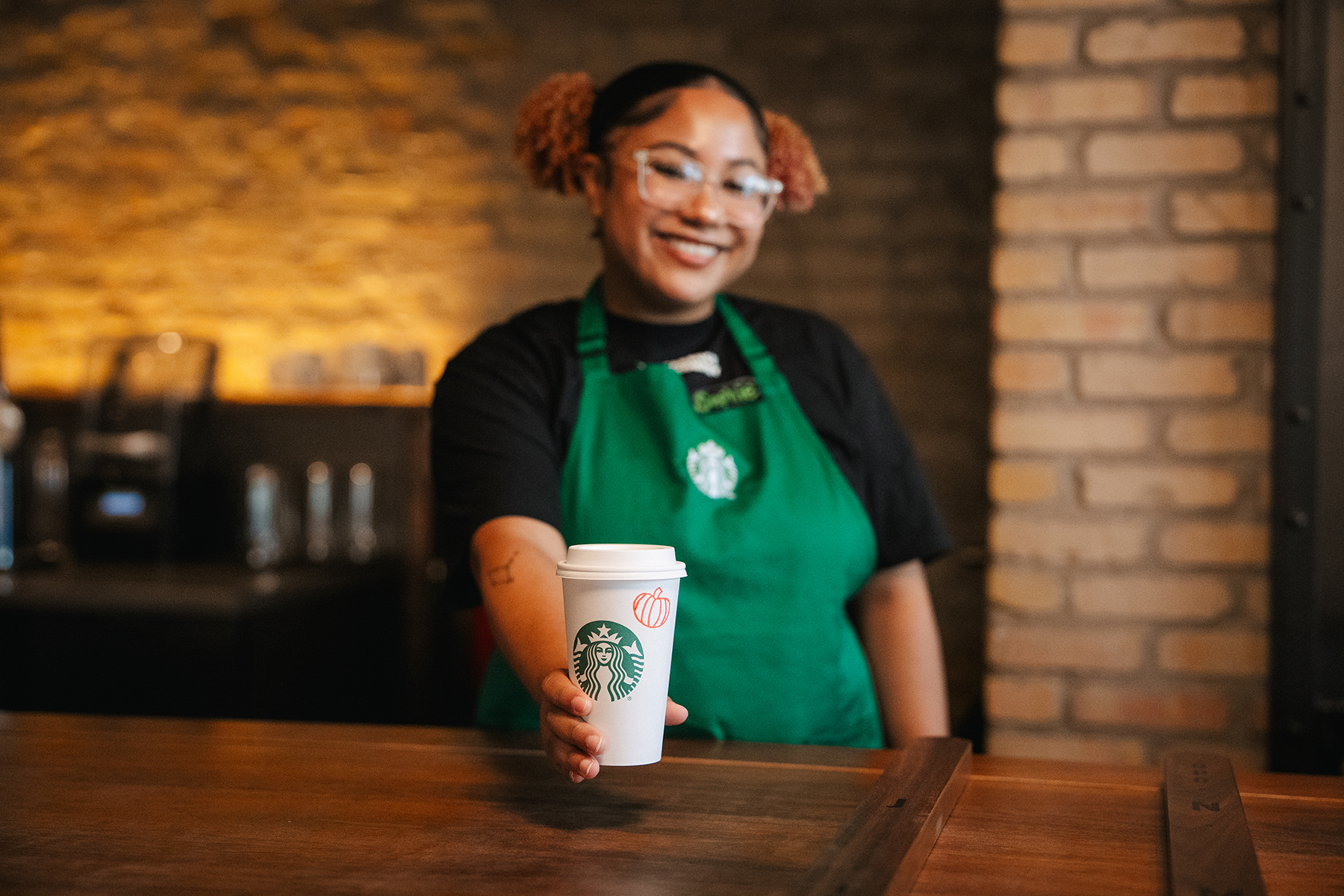 A barista in a green apron holds a Starbucks cup with a pumpkin drawing, smiling in a cozy café setting with brick walls.
