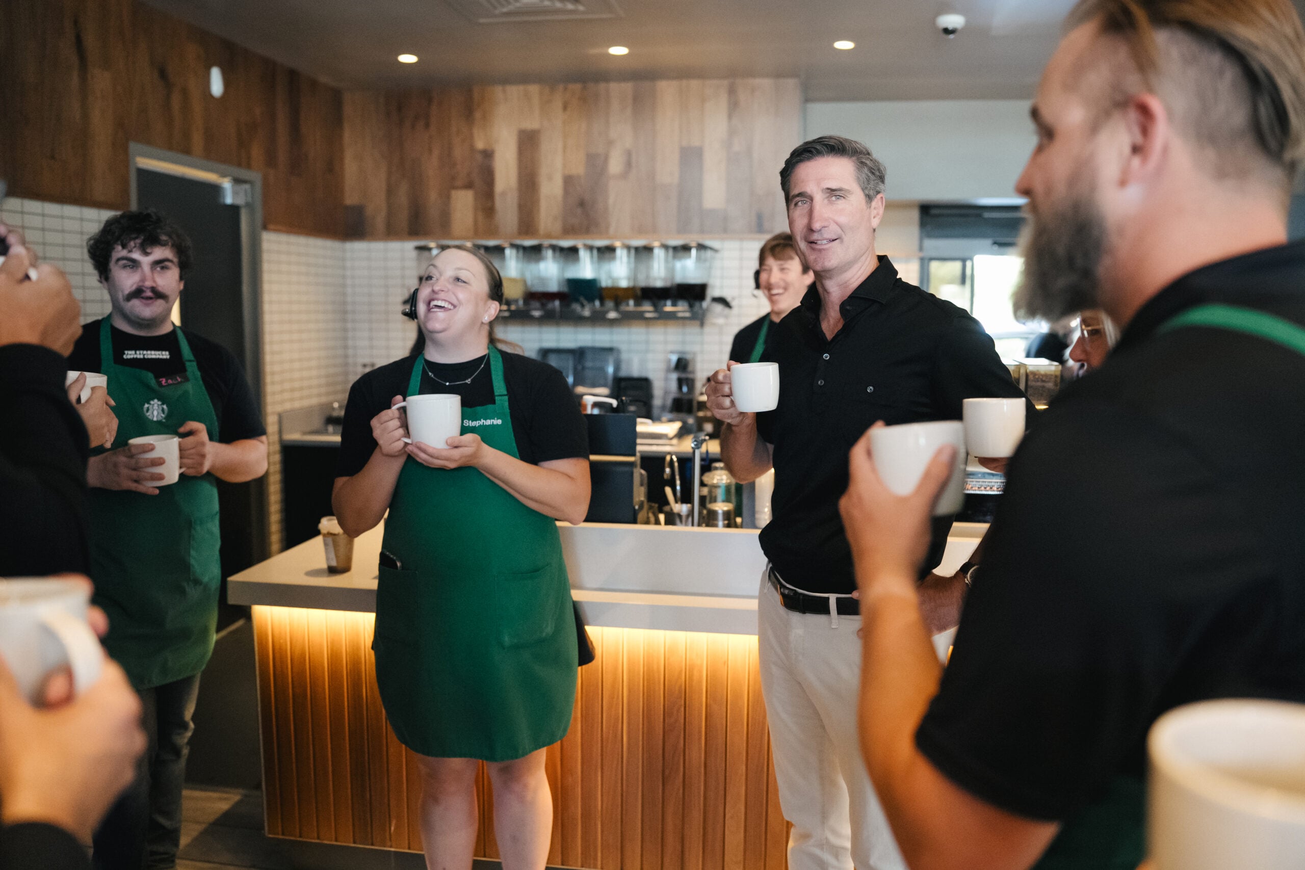 A group of people in a coffee shop, smiling and holding white mugs. One person wears a green apron, while others are casually dressed. The interior features wooden accents and a coffee bar in the background.