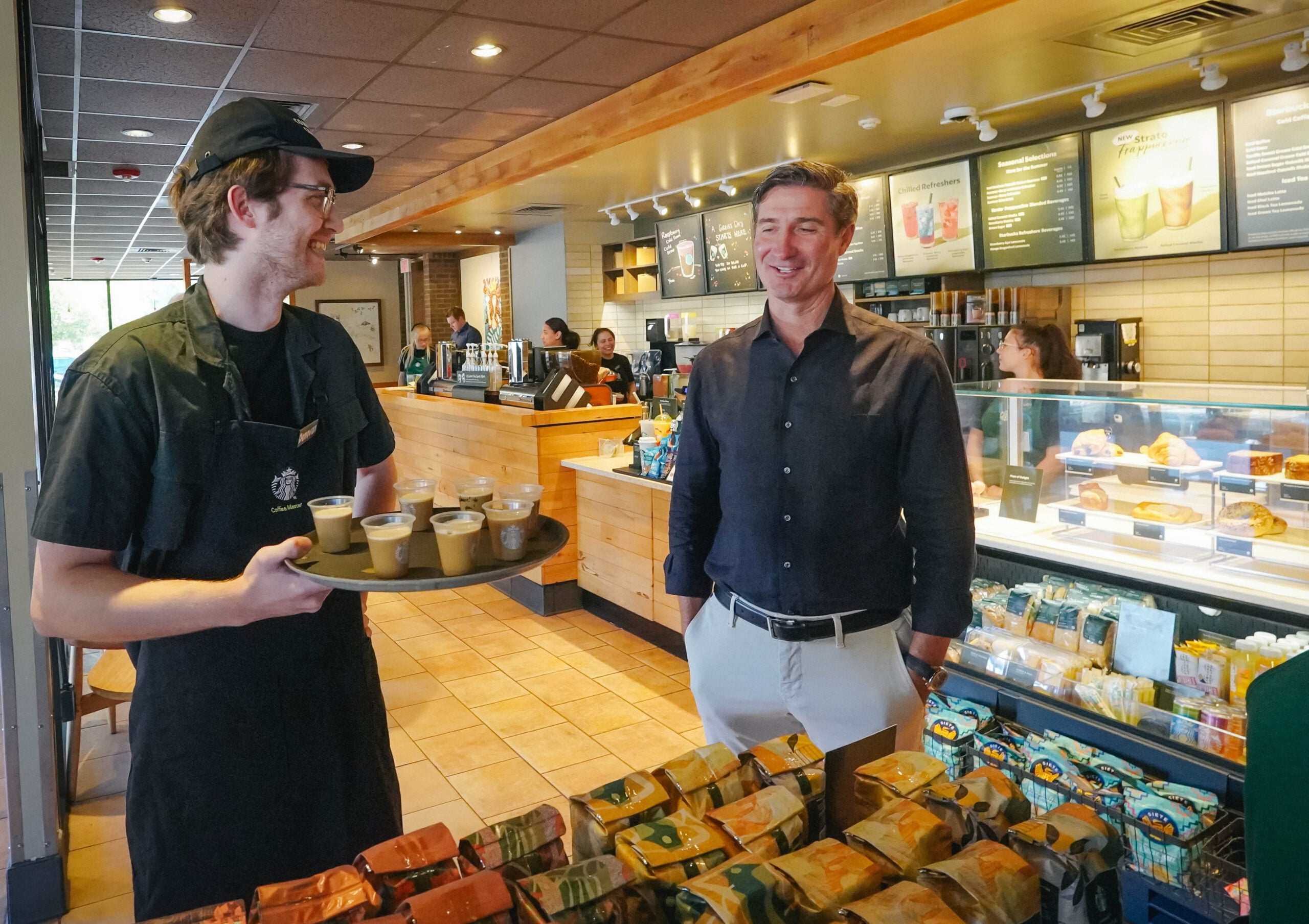 A barista in a black apron holds a tray with several cups of coffee, smiling at a man in a dark shirt. The café has wooden decor and various food items displayed.