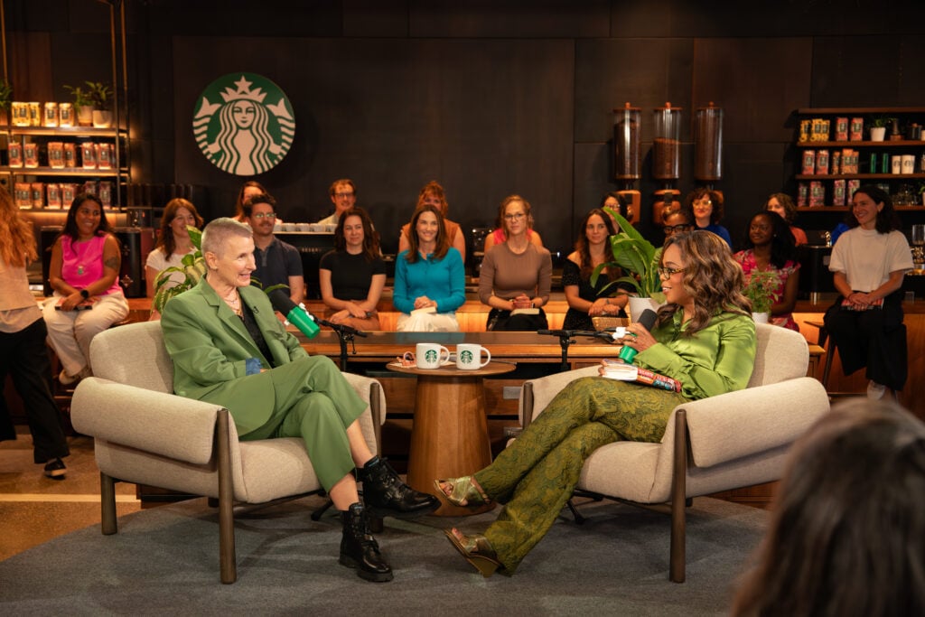 Two women are seated in armchairs, engaged in conversation. One wears a green suit and holds a drink, while the other is in a green outfit with patterned pants. A group of people is seated in the background, and there are Starbucks logos visible.