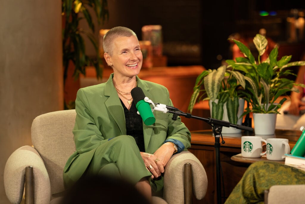 A smiling woman with short hair is seated in a light green suit, holding a microphone with a green cover. She is in a cozy setting with plants and a Starbucks cup nearby.