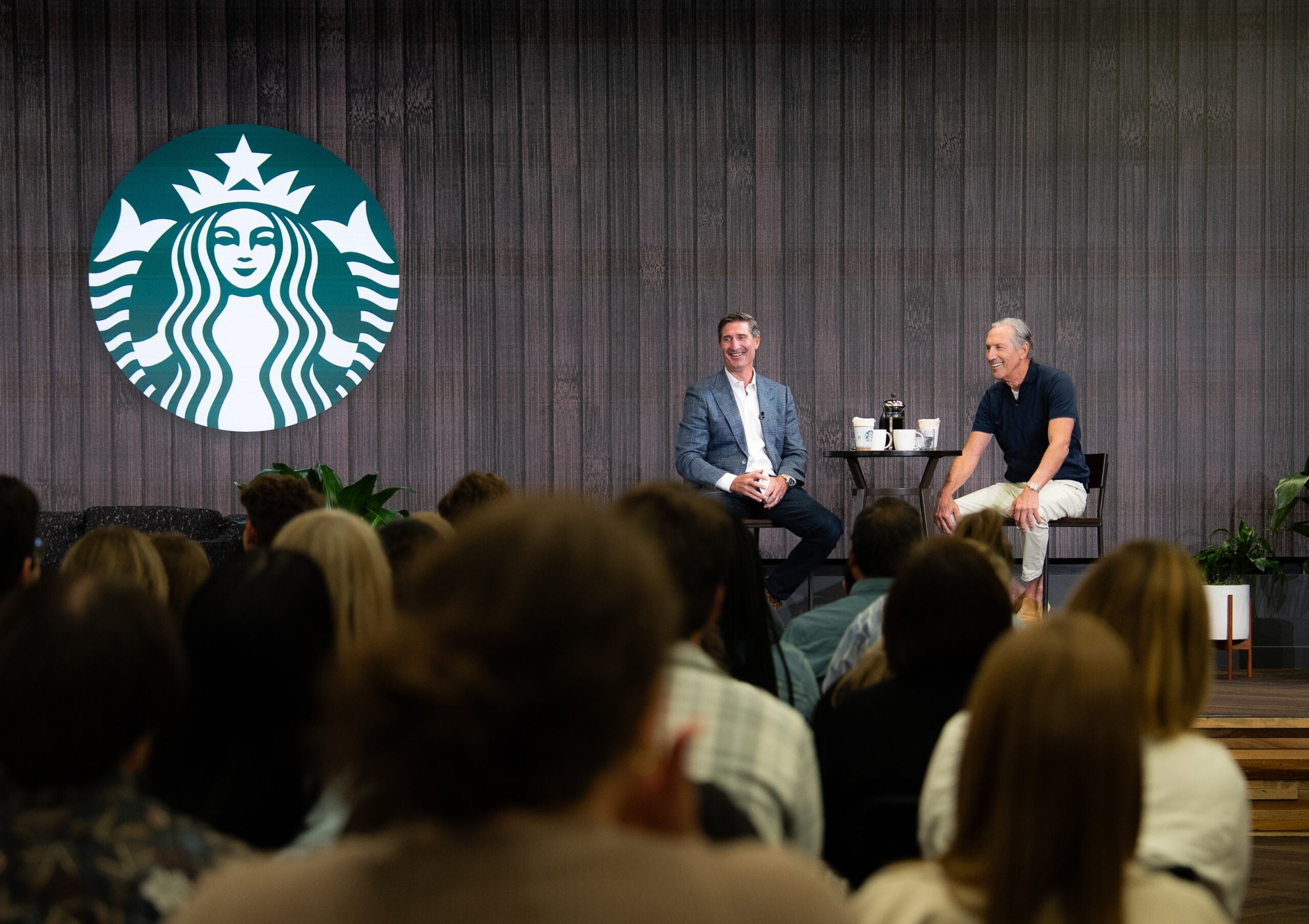Two men are seated on stage, smiling and engaging with an audience. Behind them is a large Starbucks logo on a wooden backdrop. A small table with coffee cups is positioned between them. The audience is partially visible, focusing on the speakers.