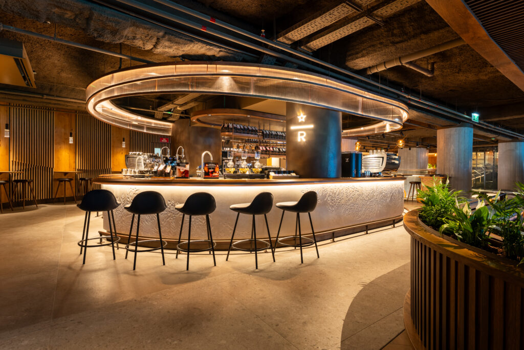 A modern bar with a sleek, circular design features a polished countertop and stylish black stools. The backdrop includes shelves with bottles and a coffee machine, illuminated by warm lighting. Greenery adds a touch of nature to the space.