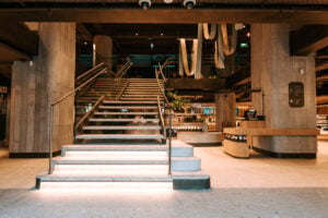 A modern interior featuring a wide staircase with wooden steps and a sleek railing. The space has warm lighting, plants, and a coffee bar in the background.