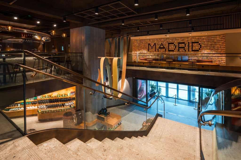 A modern interior featuring a staircase with a glass railing, leading to a lower level. The space includes wooden accents, decorative lighting, and a vibrant wall display. A checkout area is visible, along with shelves of products. Large windows allow natural light to enter.