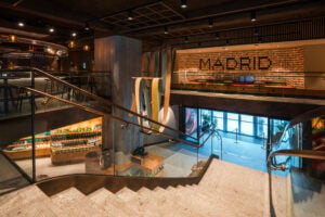 A modern interior featuring a staircase with a glass railing, leading to a lower level. The space includes wooden accents, decorative lighting, and a vibrant wall display. A checkout area is visible, along with shelves of products. Large windows allow natural light to enter.