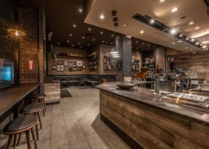 A cozy café interior featuring a wooden bar, seating area with dark booths, and a coffee counter with equipment. The walls display framed photos and art.