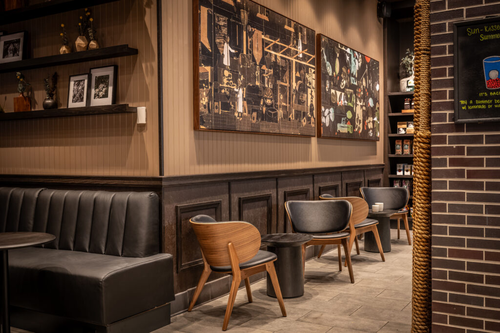 A cozy café interior featuring a black leather bench, wooden and leather chairs, and a small round table. Two large framed artworks adorn the wall, and shelves display various items. The color palette is warm and inviting.