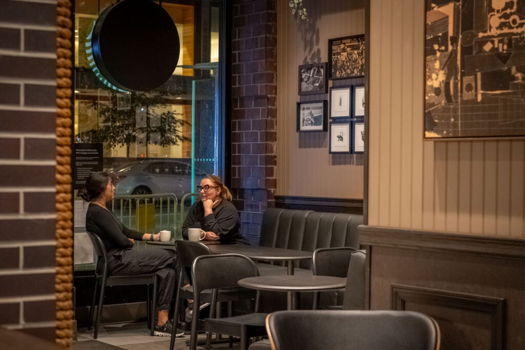 Two women sit at a table in a cozy café, engaged in conversation over cups. The interior features a mix of brick and framed artwork, with soft lighting creating a warm atmosphere.