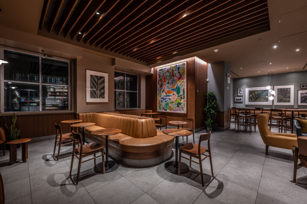 A modern café interior featuring a curved leather bench, wooden tables, and chairs. Colorful artwork adorns the wall, complemented by warm lighting and a sleek ceiling design.