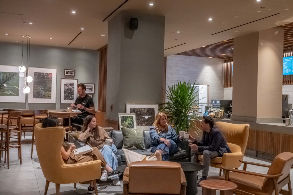 A cozy lounge area features a group of people seated on a sofa and armchairs, engaged in conversation. There are plants and framed artworks on the walls, creating a relaxed atmosphere.