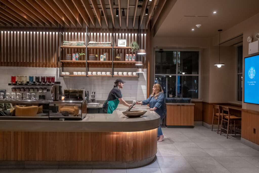A barista in a black shirt and green apron hands a cup to a woman in a blue shirt at a modern coffee shop. The interior features wooden shelves with mugs and decorative items, and soft lighting creates a warm atmosphere.