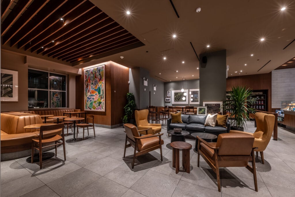 A modern lounge area featuring a mix of seating, including leather chairs and a dark sofa with decorative pillows. The space has wooden accents, a colorful wall art piece, and plants, creating a cozy atmosphere.