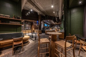 A modern café interior featuring wooden furniture, including round and square tables, and high-backed chairs. The space has a warm color palette with dark walls and wooden accents. Shelves display items and framed photos, while a coffee counter is visible in the background, illuminated by soft lighting.