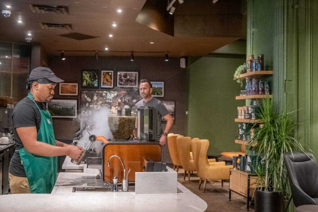 A barista in a green shirt operates a coffee machine, with steam rising from it. In the background, a man stands near yellow chairs and a shelf filled with products. Framed photos decorate the wall.