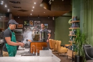 A barista in a green shirt operates a coffee machine, with steam rising from it. In the background, a man stands near yellow chairs and a shelf filled with products. Framed photos decorate the wall.