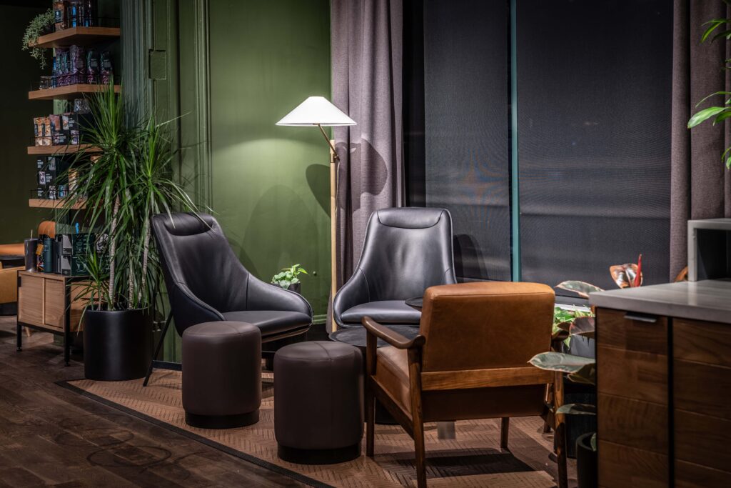 A cozy seating area featuring two modern black lounge chairs, a brown wooden armchair, and round ottomans. A floor lamp and plants add warmth.