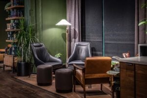 A cozy seating area featuring two modern black lounge chairs, a brown wooden armchair, and round ottomans. A floor lamp and plants add warmth.