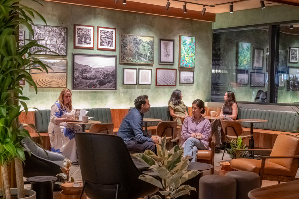 A cozy café scene featuring several people engaged in conversation. The walls are adorned with various framed artworks, and there are comfortable seating arrangements with plants and wooden tables.