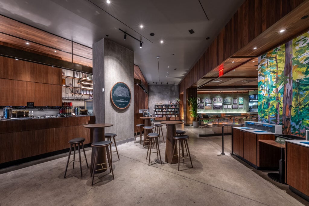 A modern café interior featuring wooden accents, high-top tables, and stools. A bar area is visible with shelves and a display of products. The space is well-lit with a mix of natural and artificial lighting.