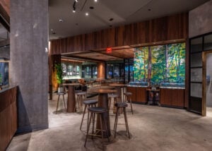 A modern café interior featuring wooden accents, high tables, and stools. A vibrant mural of trees adorns one wall, while glass displays and seating areas are visible in the background.