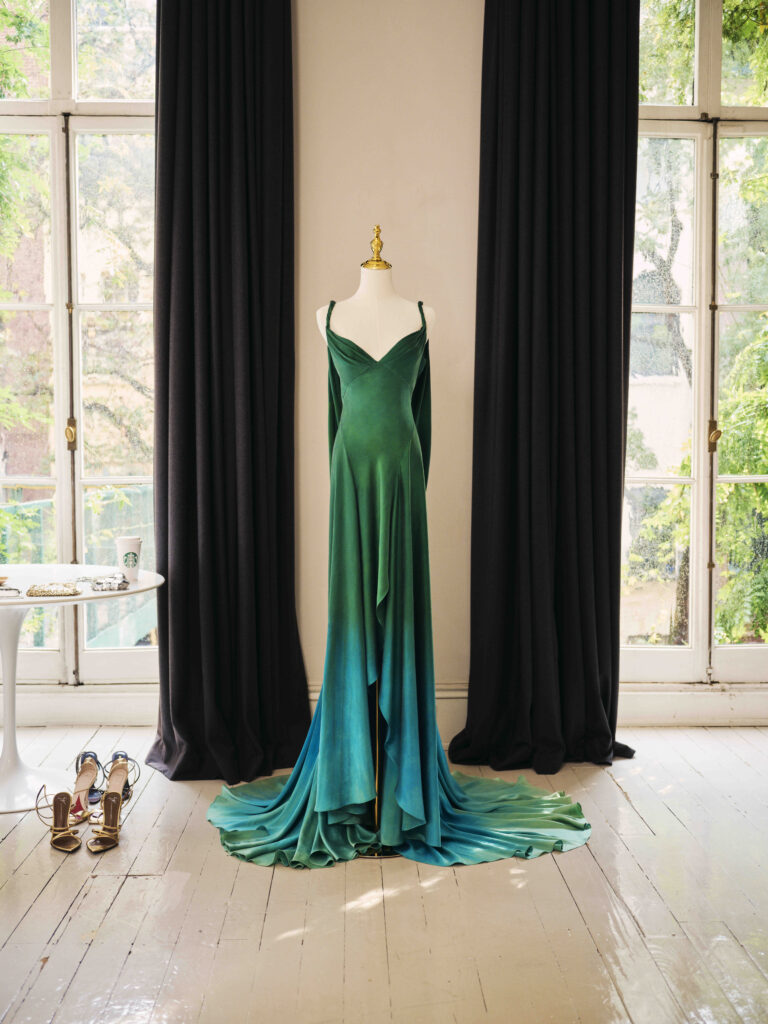 A flowing green dress on a mannequin, featuring a deep V-neck and long draped fabric. Nearby, a small table holds accessories and shoes.