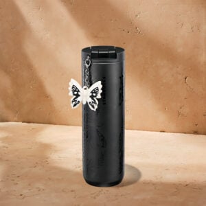 A sleek black Starbucks travel mug features a butterfly charm attached to its side, set against a textured beige background.